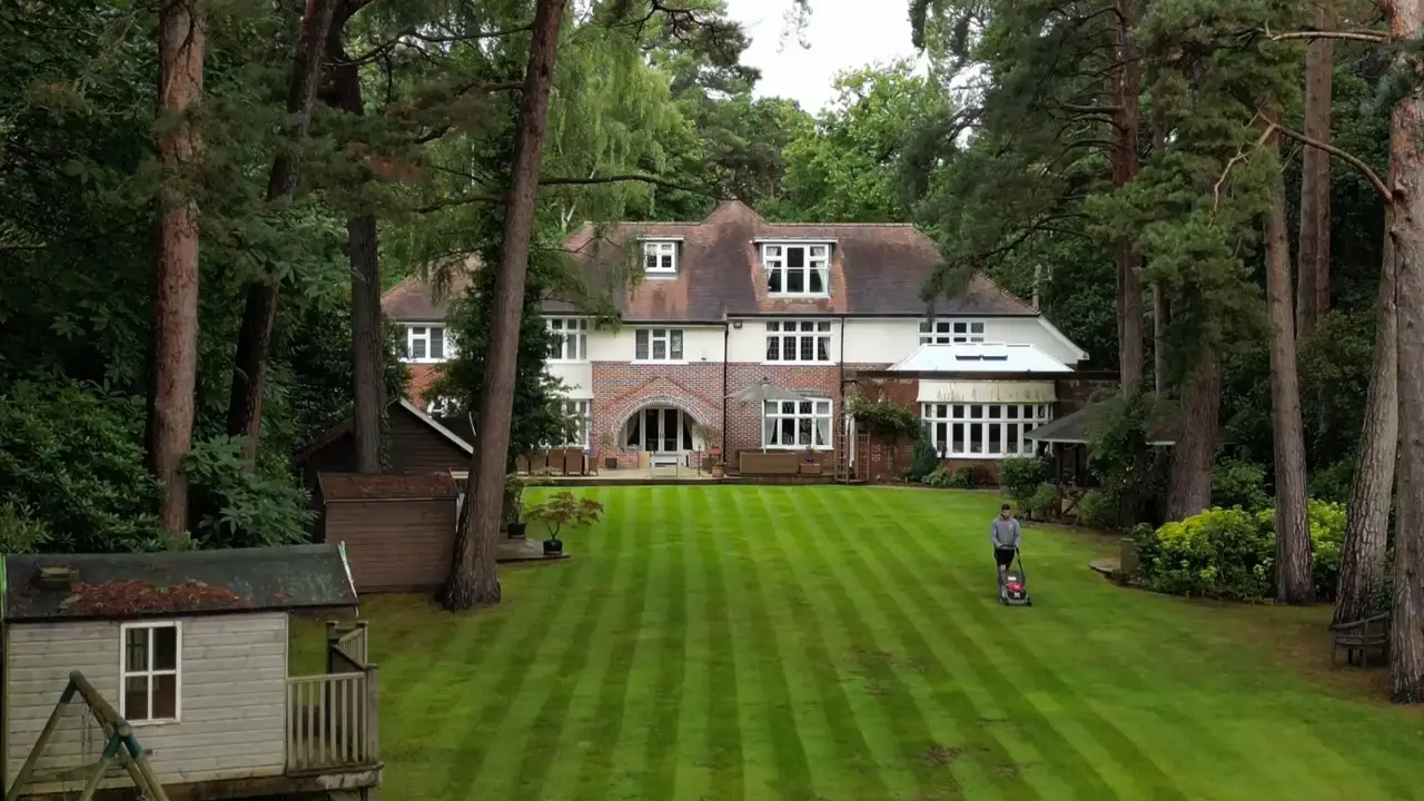 A large house in wooded grounds with a striped lawn being mown